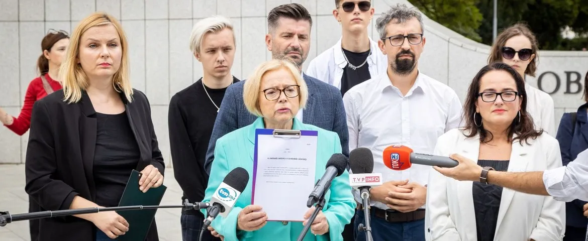 Na zdjęciu grupa osób stoi przed kamerami, z tą, która trzyma dokument, wyróżniającą się w centralnej części kadru.