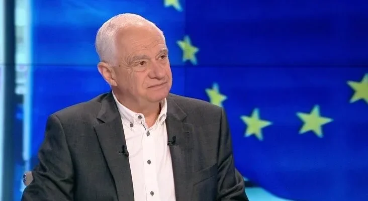 Mężczyzna w marynarce i białej koszuli siedzi w studiu telewizyjnym na tle flagi Unii Europejskiej.