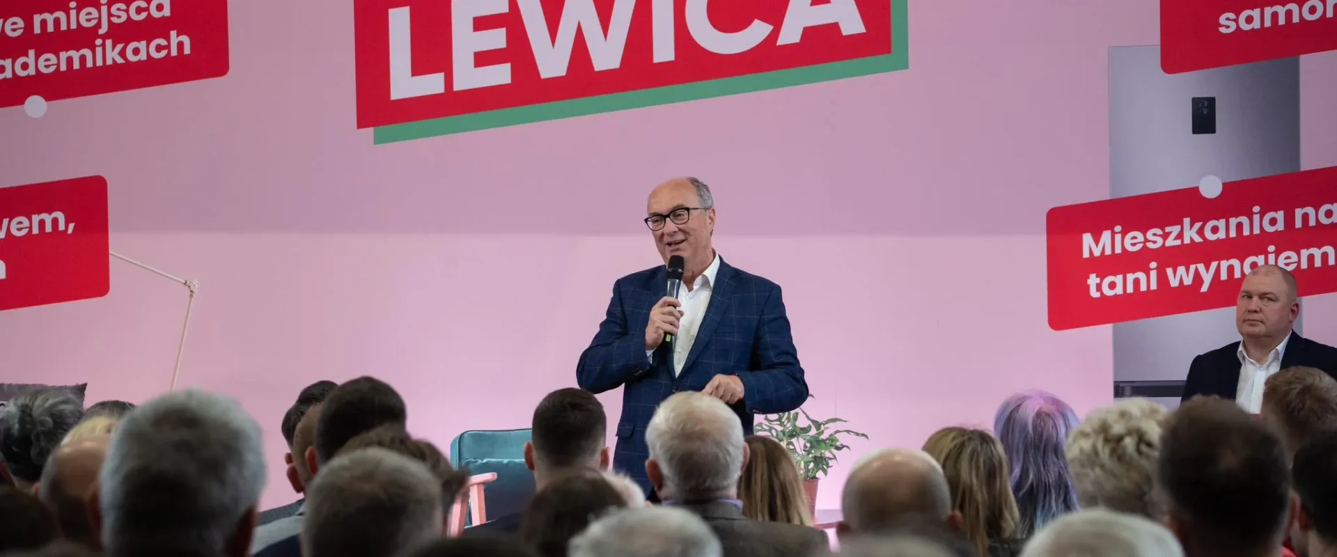 Polityk Lewicy w garniturze przemawia do zgromadzonej publiczności na scenie z różową scenografią i dużym napisem LEWICA w tle.