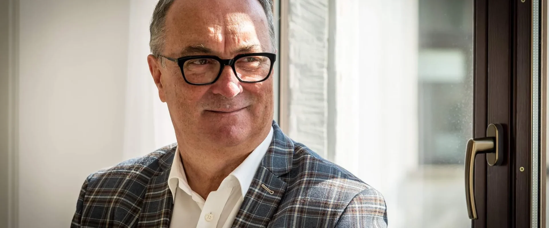 Mężczyzna w okularach, w kraciastym garniturze i białej koszuli, siedzi przy oknie i uśmiecha się.