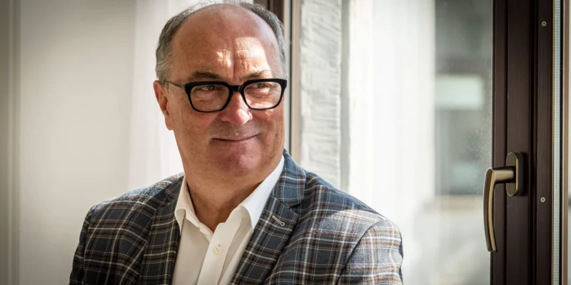 Mężczyzna w okularach, w kraciastym garniturze i białej koszuli, siedzi przy oknie i uśmiecha się.