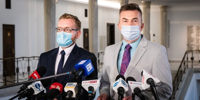 Dwóch mężczyzn w garniturach i maseczkach udziela konferencji prasowej w budynku rządowym, stojąc przed licznymi mikrofonami, w kontekście wniosku o informację Prezesa Rady Ministrów dotyczącego wykorzystania Funduszu Sprawiedliwości w latach 2018–2020.