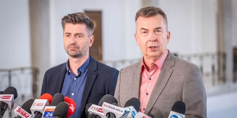 Dwóch mężczyzn w marynarkach stoi przy licznych mikrofonach na konferencji prasowej w jasnym wnętrzu.