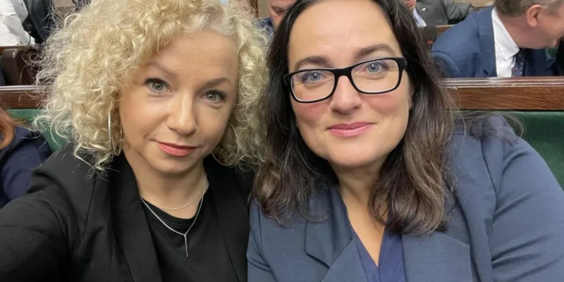 Na zdjęciu dwie kobiety siedzą obok siebie w sali, uśmiechając się do kamery, w tle widać innych uczestników spotkania.