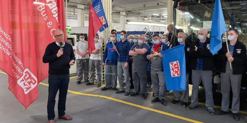 Na zdjęciu przedstawiono grupę pracowników przemysłowych, którzy uczestniczą w wydarzeniu, gdzie jeden z mężczyzn przemawia, trzymając mikrofon, a w tle widoczne są flagi oraz maszyny.