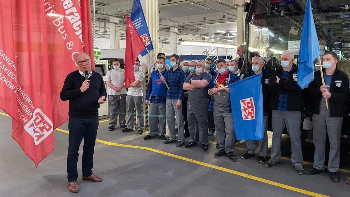 Na zdjęciu przedstawiono grupę pracowników przemysłowych, którzy uczestniczą w wydarzeniu, gdzie jeden z mężczyzn przemawia, trzymając mikrofon, a w tle widoczne są flagi oraz maszyny.