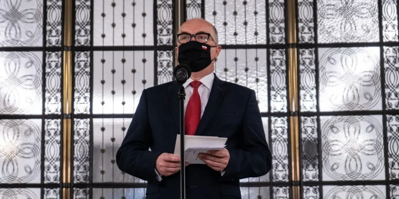 Mężczyzna w garniturze i czerwonym krawacie stoi przy mikrofonie, trzyma kartki, ma czarną maseczkę z polską flagą i stoi przed ozdobnymi metalowymi drzwiami.