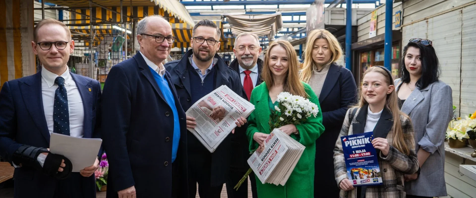 Grupa osób stojących na targowisku pozuje do zdjęcia; kobieta w zielonym płaszczu trzyma bukiet kwiatów i gazetę, inni mężczyźni trzymają gazety.