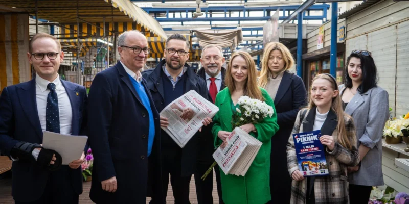 Grupa osób stojących na targowisku pozuje do zdjęcia; kobieta w zielonym płaszczu trzyma bukiet kwiatów i gazetę, inni mężczyźni trzymają gazety.