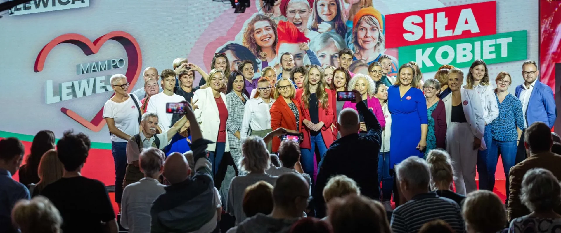 Grupa kobiet na scenie podczas wydarzenia Siła Kobiet, pozująca do wspólnego zdjęcia na tle kolorowego baneru z napisem Siła Kobiet i zgromadzonego tłumu widzów.