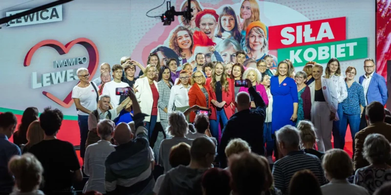 Grupa kobiet na scenie podczas wydarzenia Siła Kobiet, pozująca do wspólnego zdjęcia na tle kolorowego baneru z napisem Siła Kobiet i zgromadzonego tłumu widzów.