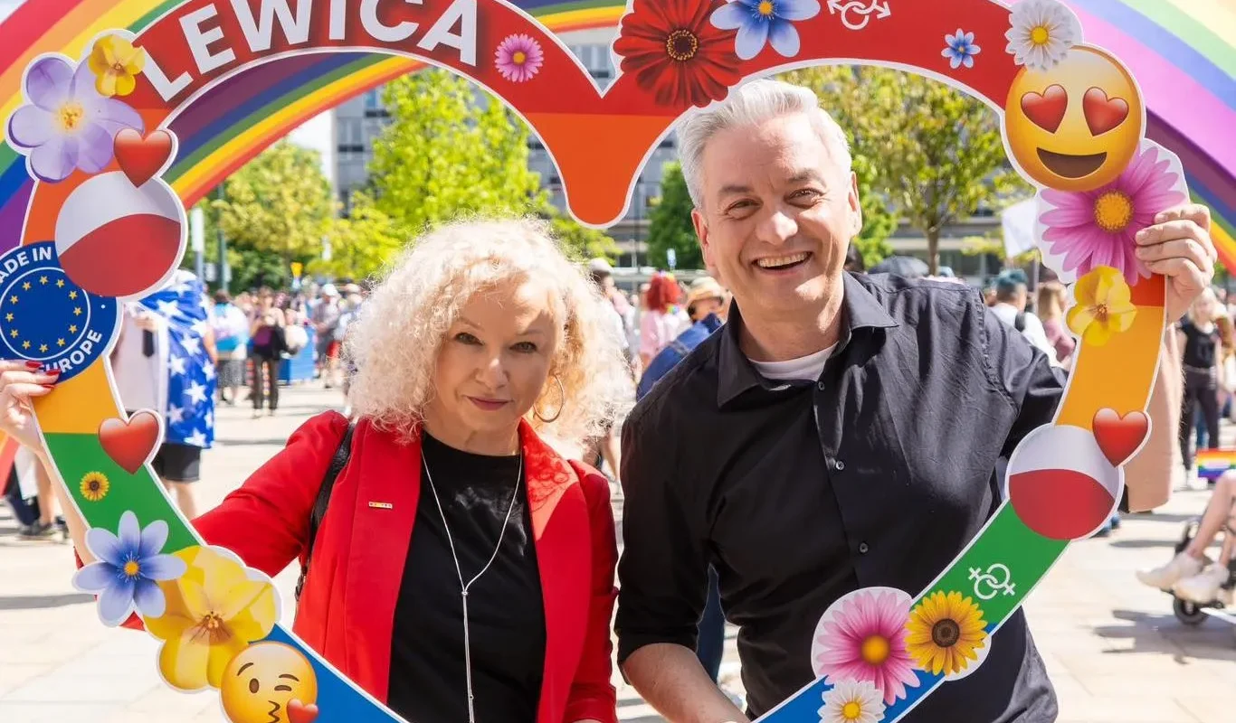 Dwoje uśmiechniętych ludzi stoi na plenerowym wydarzeniu, trzymając kolorową, tęczową ramkę w kształcie serca z napisem LEWICA i kwiatowymi dekoracjami.