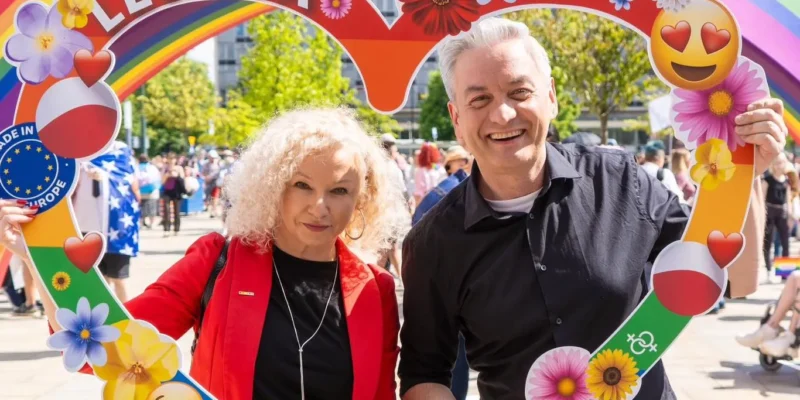 Dwoje uśmiechniętych ludzi stoi na plenerowym wydarzeniu, trzymając kolorową, tęczową ramkę w kształcie serca z napisem LEWICA i kwiatowymi dekoracjami.