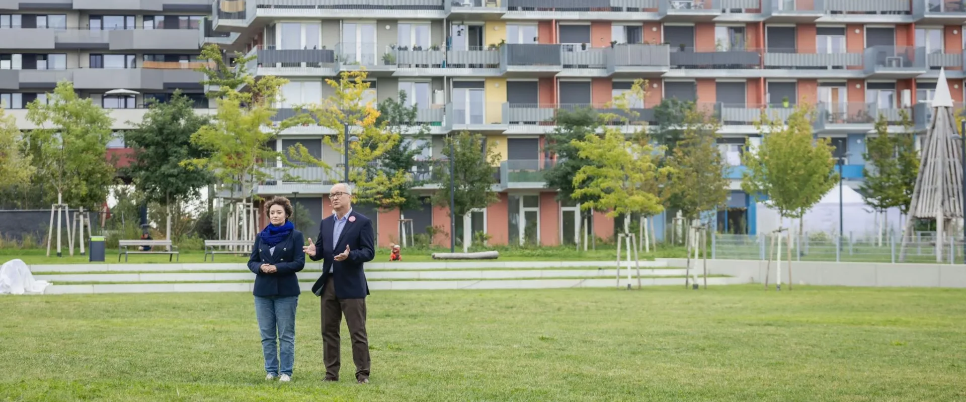 Dwoje dorosłych stoi na zielonej trawie przed kolorowym, wielopiętrowym blokiem mieszkalnym z balkonami.
