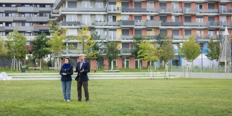 Dwoje dorosłych stoi na zielonej trawie przed kolorowym, wielopiętrowym blokiem mieszkalnym z balkonami.