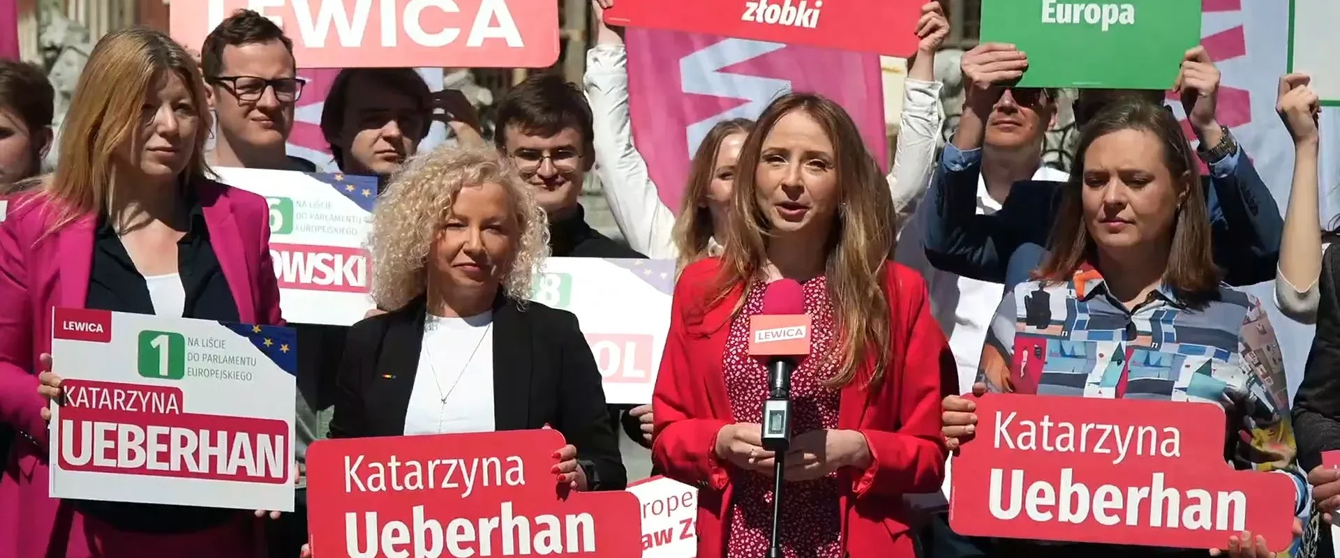 Grupa demonstrantów Lewicy z plakatami promującymi work‑life balance i dostęp do żłobków, a kobieta w czerwonym żakiecie przemawia do mikrofonu.