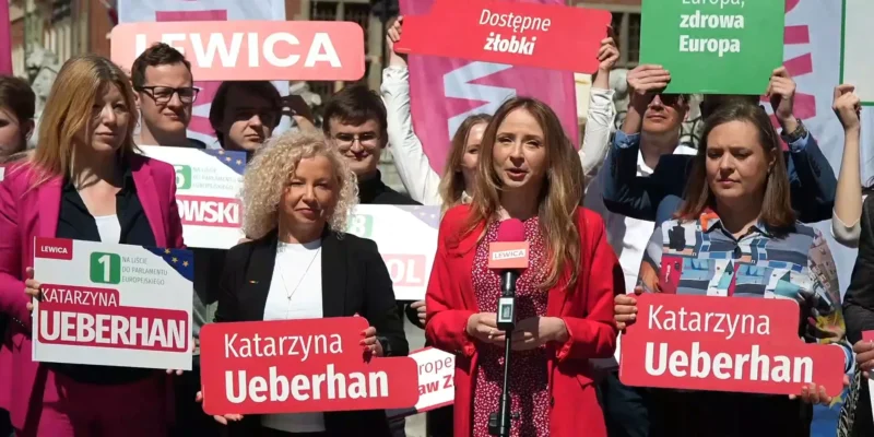 Grupa demonstrantów Lewicy z plakatami promującymi work‑life balance i dostęp do żłobków, a kobieta w czerwonym żakiecie przemawia do mikrofonu.