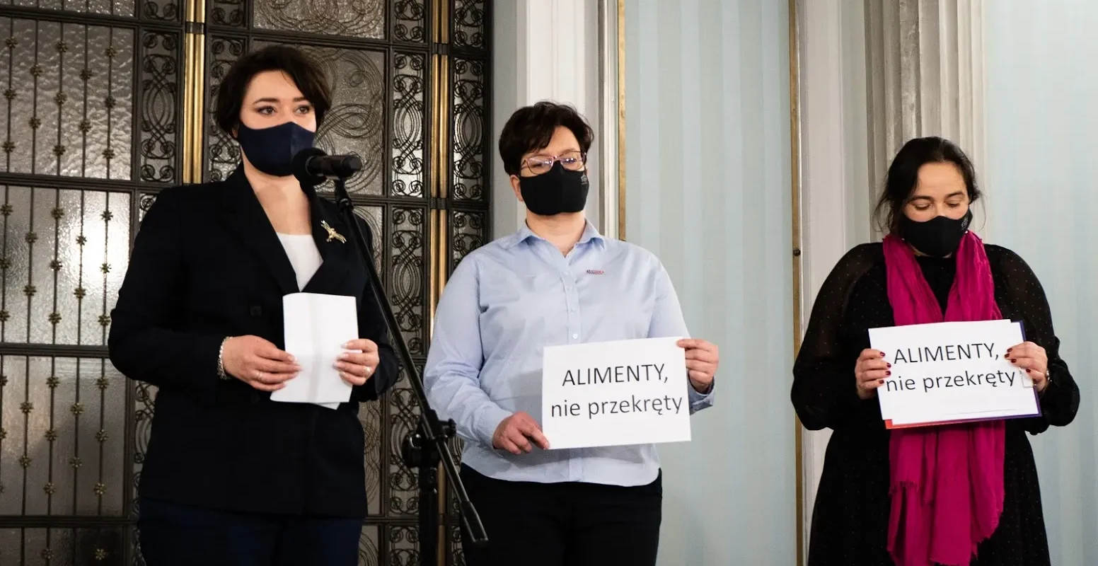 Trzy osoby w maseczkach stoją na konferencji prasowej, jedna przy mikrofonie, dwie trzymają plakaty z napisem „ALIMENTY, nie przekręty”, na tle ozdobnych drzwi i kolumn.