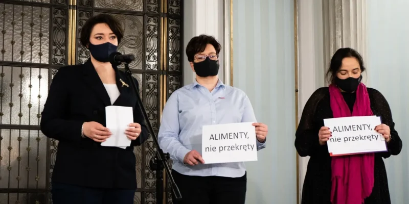 Trzy osoby w maseczkach stoją na konferencji prasowej, jedna przy mikrofonie, dwie trzymają plakaty z napisem „ALIMENTY, nie przekręty”, na tle ozdobnych drzwi i kolumn.