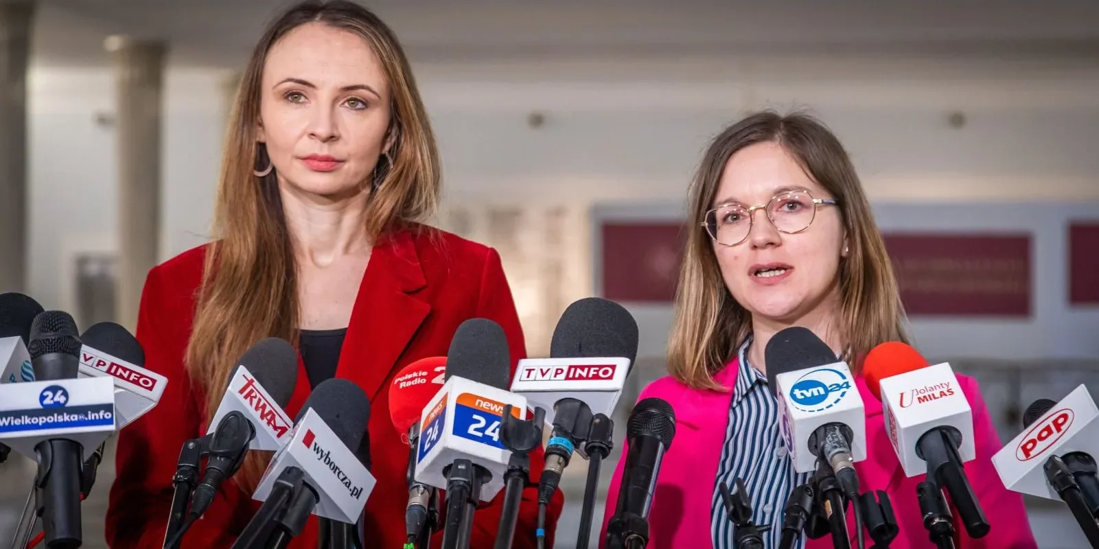 Na zdjęciu dwie kobiety stoją przy mównicy otoczonej mikrofonami, uczestnicząc w konferencji prasowej.