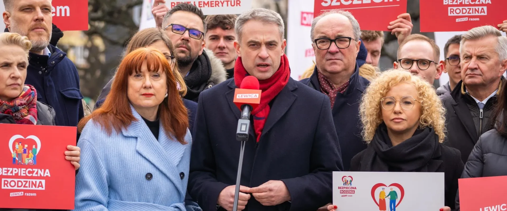 Grupa zwolenników Lewicy podczas wiecu; centralnie stojący mężczyzna w ciemnym płaszczu i czerwonym szaliku przemawia do mikrofonu, otoczony plakatami z hasłami Bezpieczna Rodzina i Szczecin.