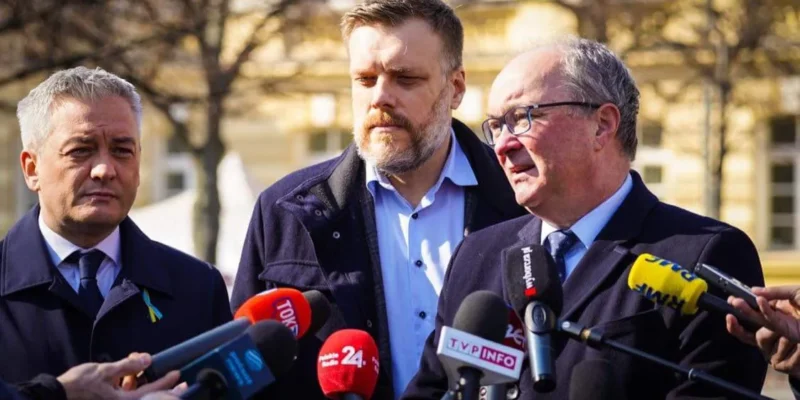 Trzech mężczyzn w garniturach stoi przed mikrofonami na konferencji prasowej w plenerze.