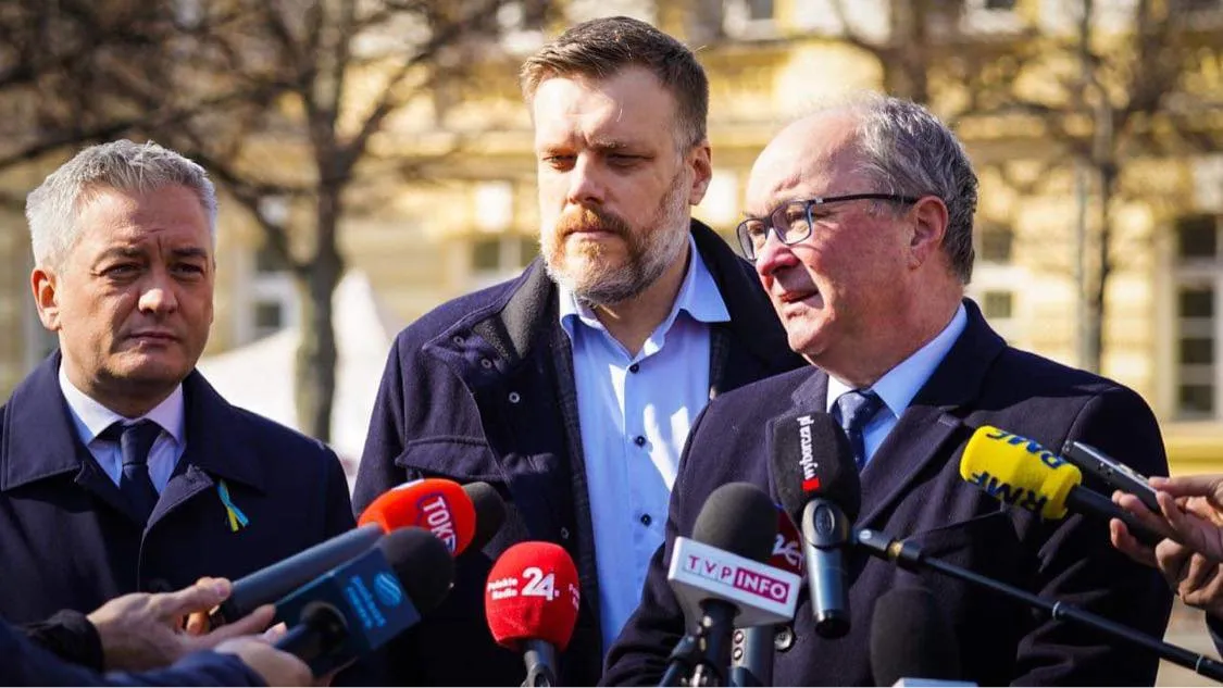 Trzech mężczyzn w garniturach stoi przed mikrofonami na konferencji prasowej w plenerze.