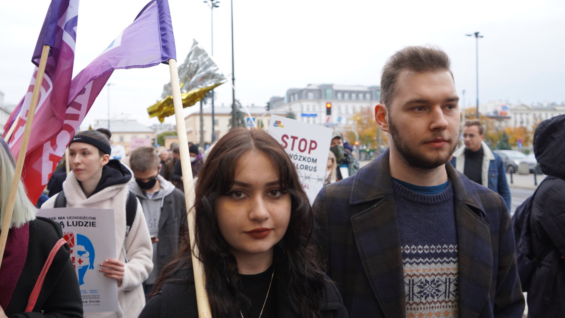 Grupa demonstrantów w marszu solidarności z migrantami z plakatami i flagami; na pierwszym planie młoda kobieta i młody mężczyzna stojący blisko siebie, w tle ulica i budynki.