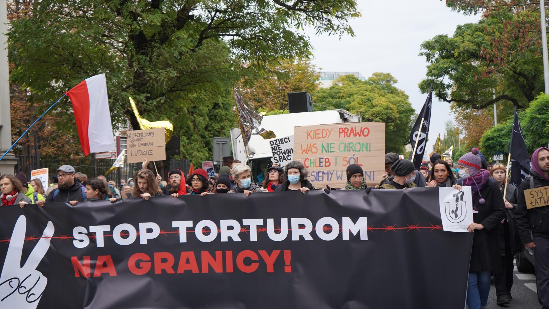 Na zdjęciu widać protestujących ludzi niosących banery z hasłami przeciwko torturom na granicy, wśród których znajdują się polska flaga oraz transparenty z różnymi hasłami społecznymi.