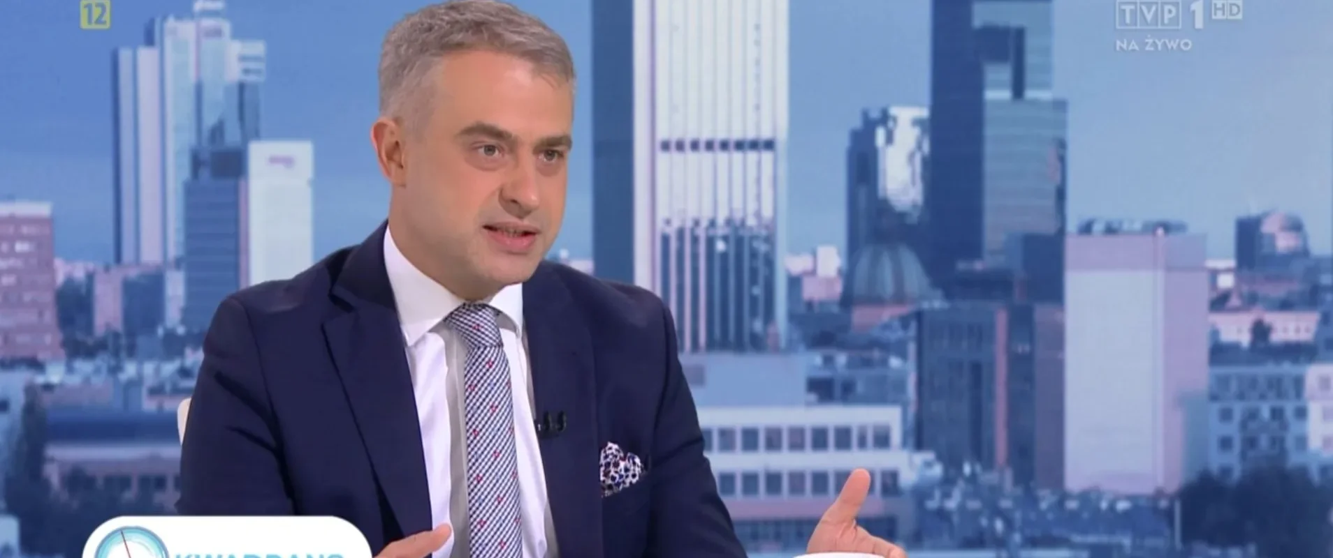 Na zdjęciu widoczny jest mężczyzna w garniturze, który rozmawia podczas programu telewizyjnego na tle miejskiego krajobrazu.