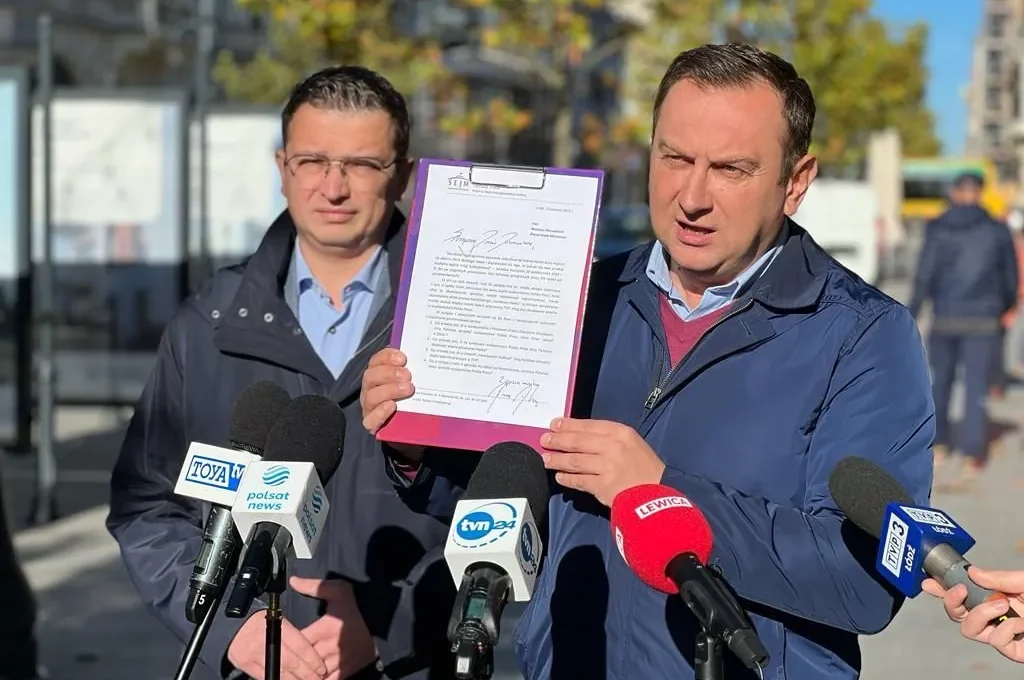 Dwóch mężczyzn na konferencji prasowej prezentuje dokument w fioletowej oprawie, stojąc przed mikrofonami mediów.