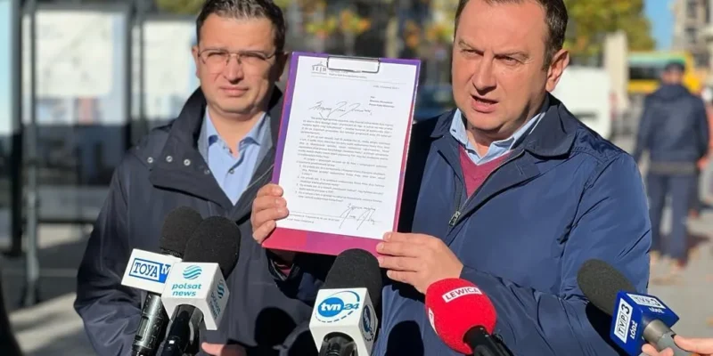 Dwóch mężczyzn na konferencji prasowej prezentuje dokument w fioletowej oprawie, stojąc przed mikrofonami mediów.