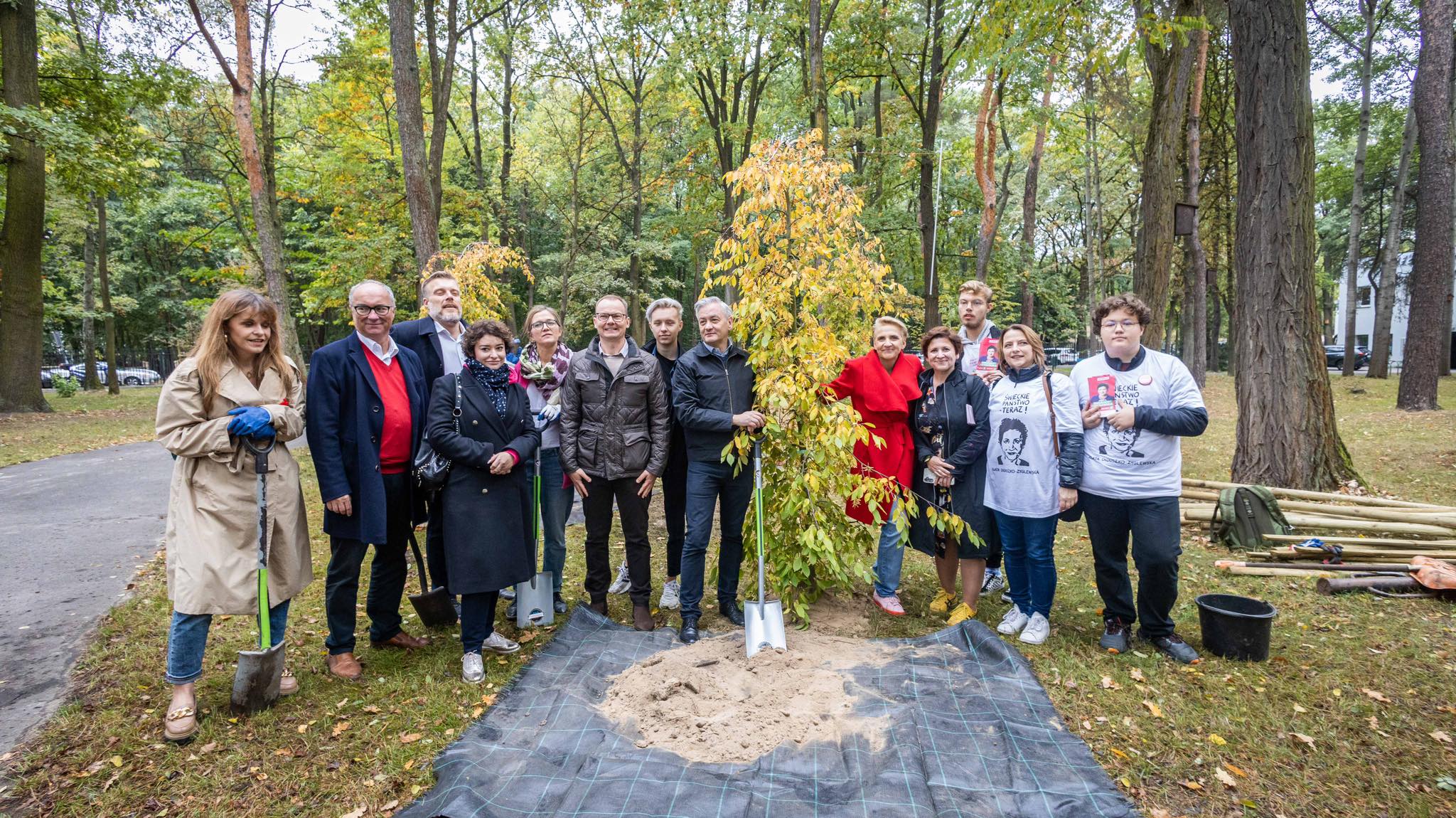 Grupa osób w parku stoi wokół młodego drzewka posadzonego na czarnej folii, z łopatami i narzędziami do sadzenia w pobliżu.
