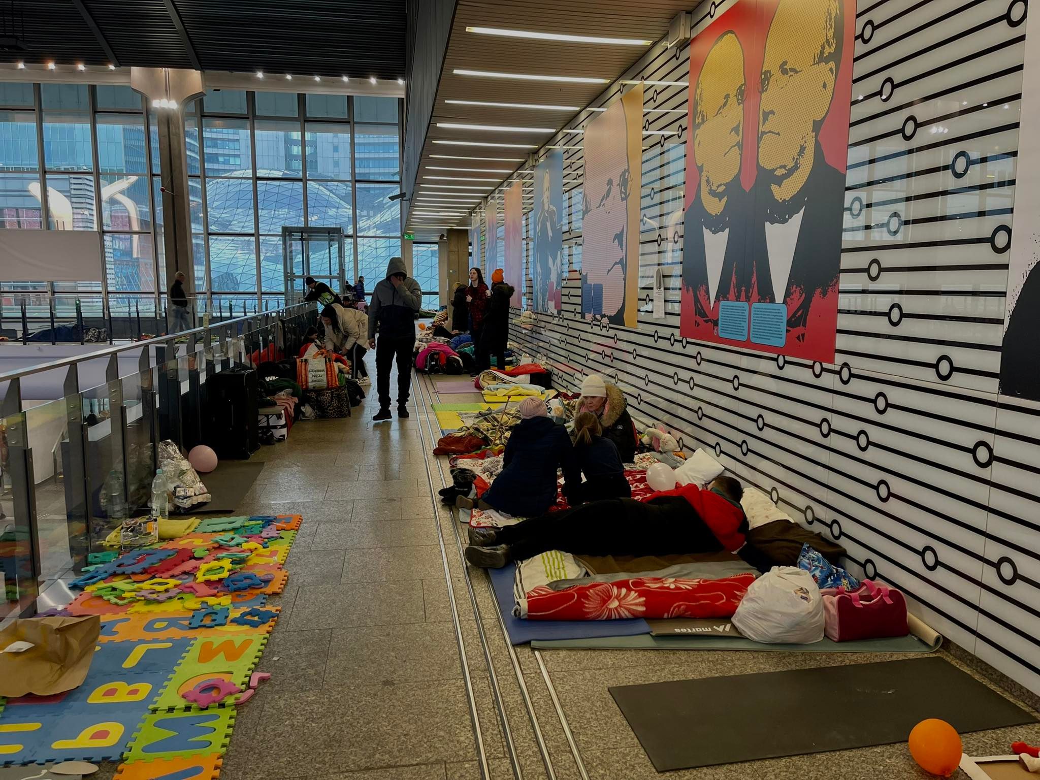 Scena na warszawskim dworcu: ludzie odpoczywają na materacach i kocach rozłożonych na podłodze, otoczeni bagażami, obok kolorowe maty dla dzieci i muralna ściana.