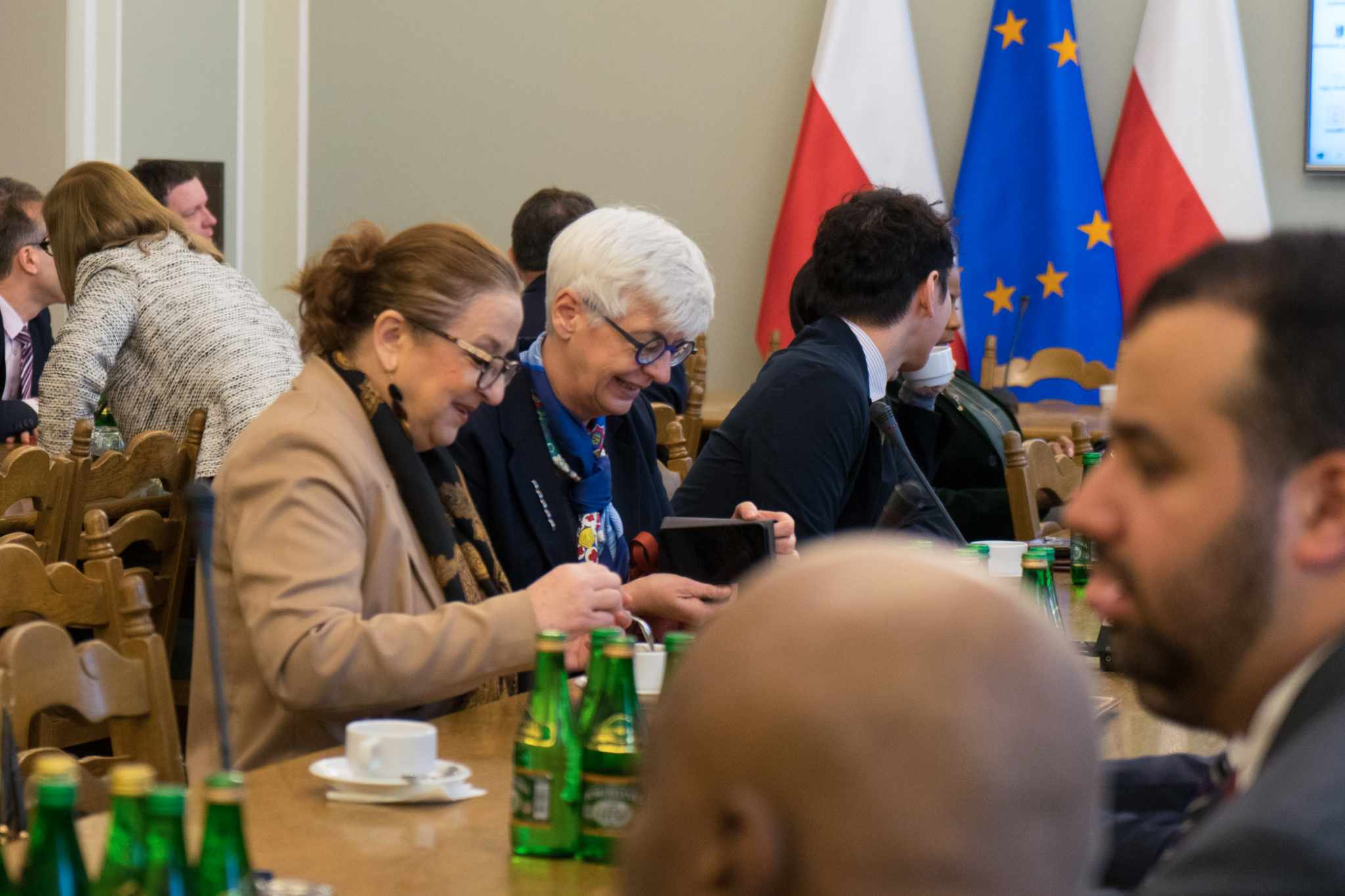 Podczas spotkania z korpusem dyplomatycznym grupa dyplomatów siedzi przy długim stole, rozmawia i przegląda materiały, a w tle widoczne są flagi Polski i Unii Europejskiej.