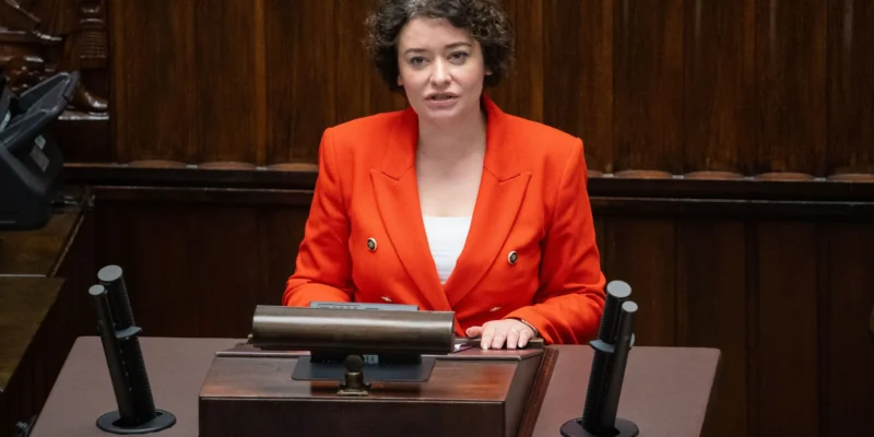 Kobieta z krótkimi, kręconymi włosami w czerwonej marynarce siedzi przy mównicy w sali plenarnej.