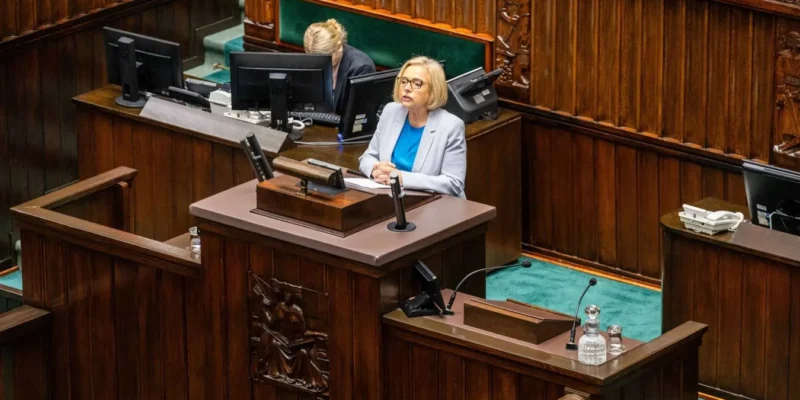 Posłanka wygłasza przemówienie z mównicy w Sejmie, w otoczeniu drewnianej architektury sali plenarnej i monitorów.