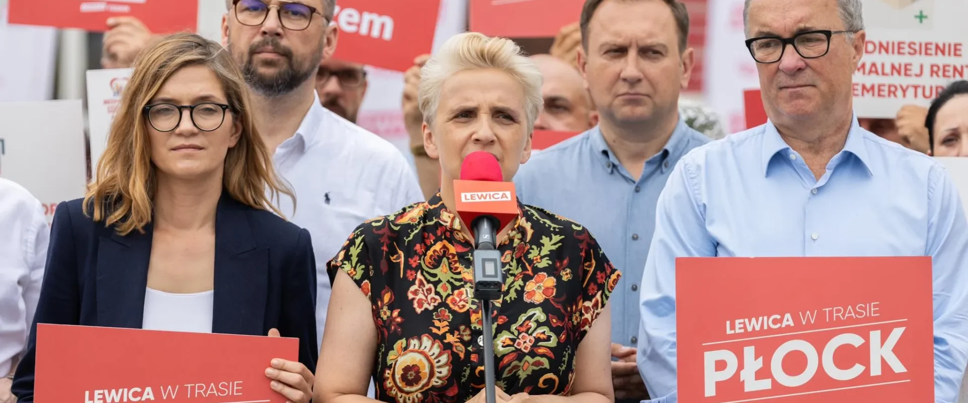 Grupa polityków Lewicy na konferencji prasowej w Płocku; centralnie przemawia do mikrofonu blondynka, otoczona innymi osobami trzymającymi czerwone plakaty z napisem PŁOCK.