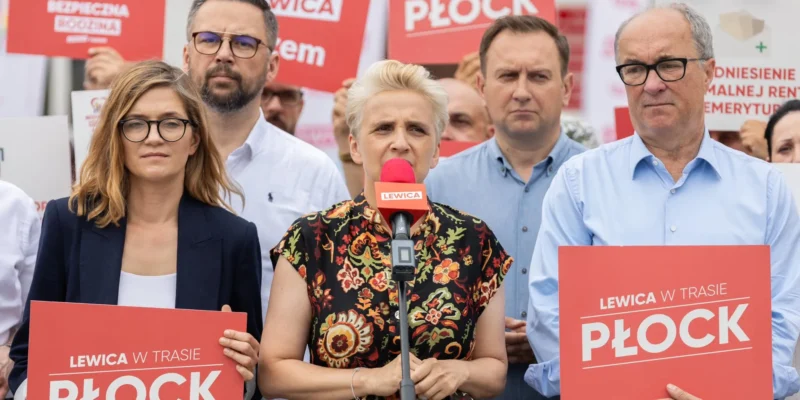 Grupa polityków Lewicy na konferencji prasowej w Płocku; centralnie przemawia do mikrofonu blondynka, otoczona innymi osobami trzymającymi czerwone plakaty z napisem PŁOCK.