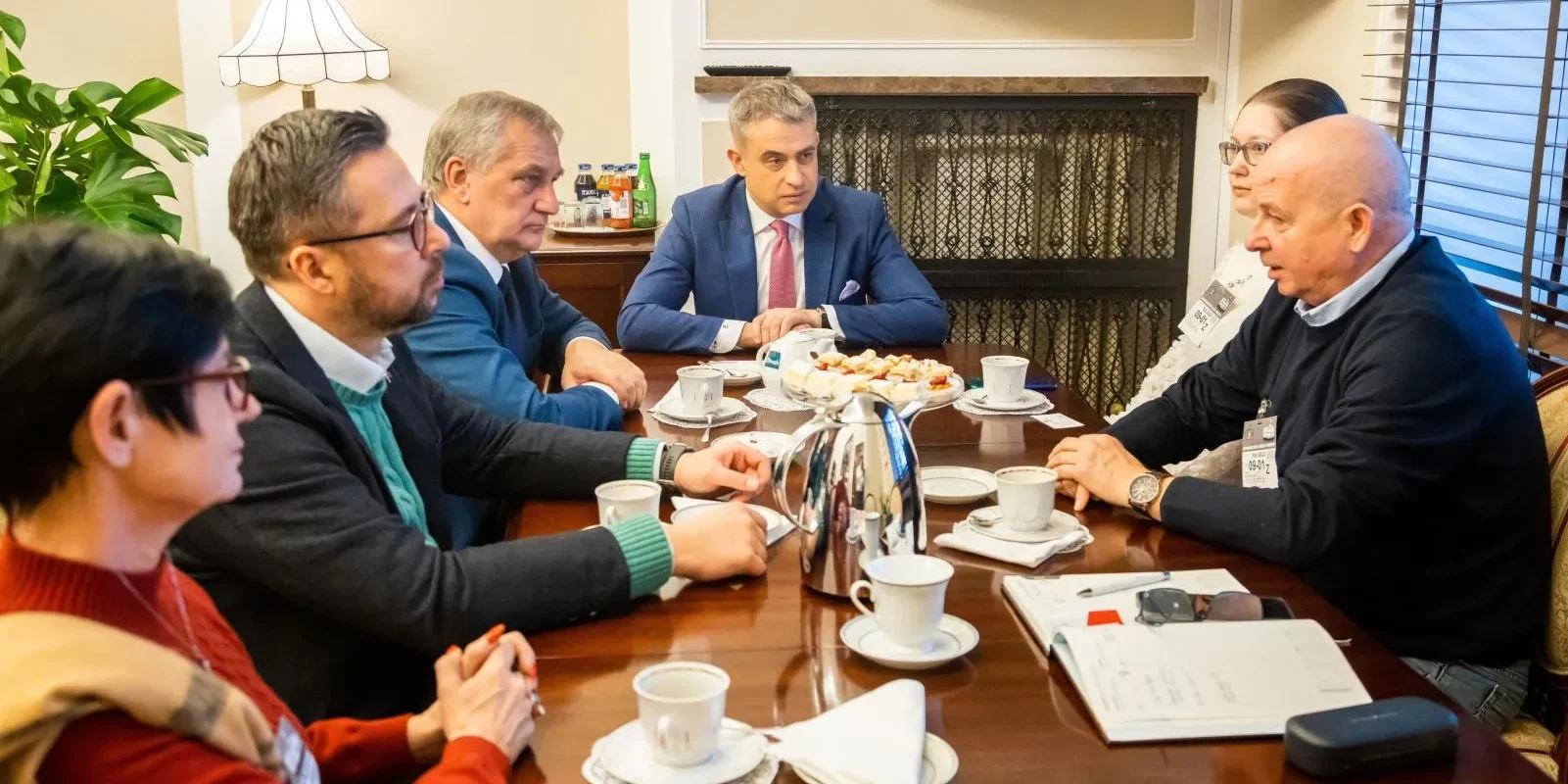 Sześciu uczestników w formalnych ubraniach siedzi przy stole podczas spotkania Bractwa Mundurowego RP, na którym ustawione są filiżanki kawy i herbaty oraz przekąski.