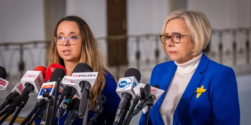 Na zdjęciu widać dwie kobiety wypowiadające się podczas konferencji prasowej, otoczone mikrofonami, w eleganckich niebieskich strojach.