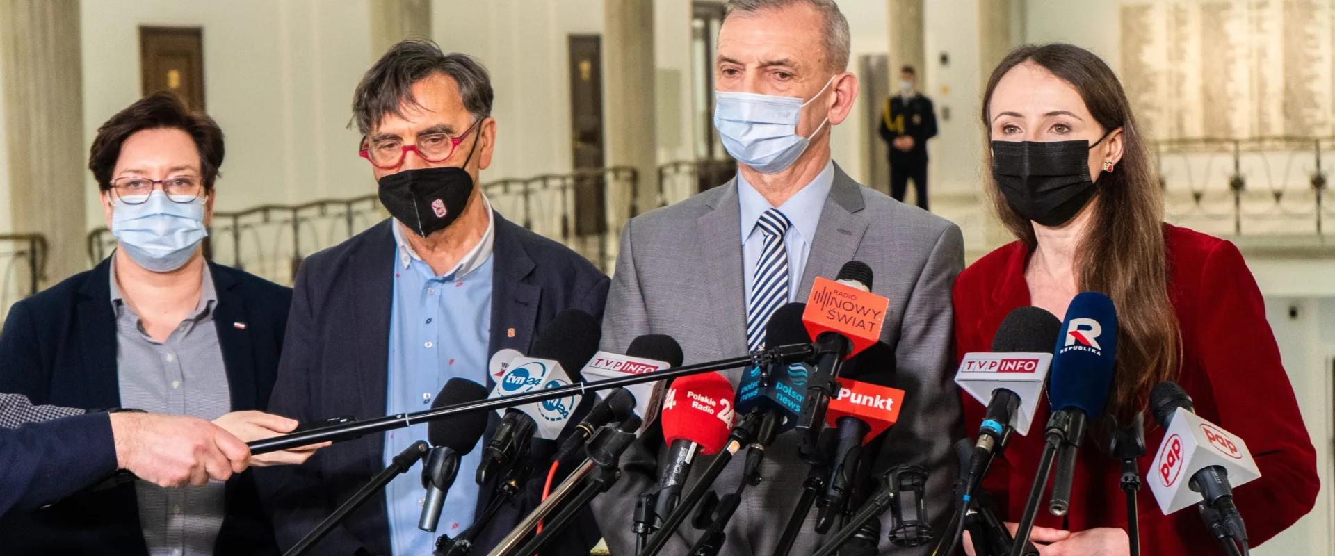 Grupa osób w maseczkach udziela konferencji prasowej, stojąc za licznymi mikrofonami ustawionymi na stojakach.