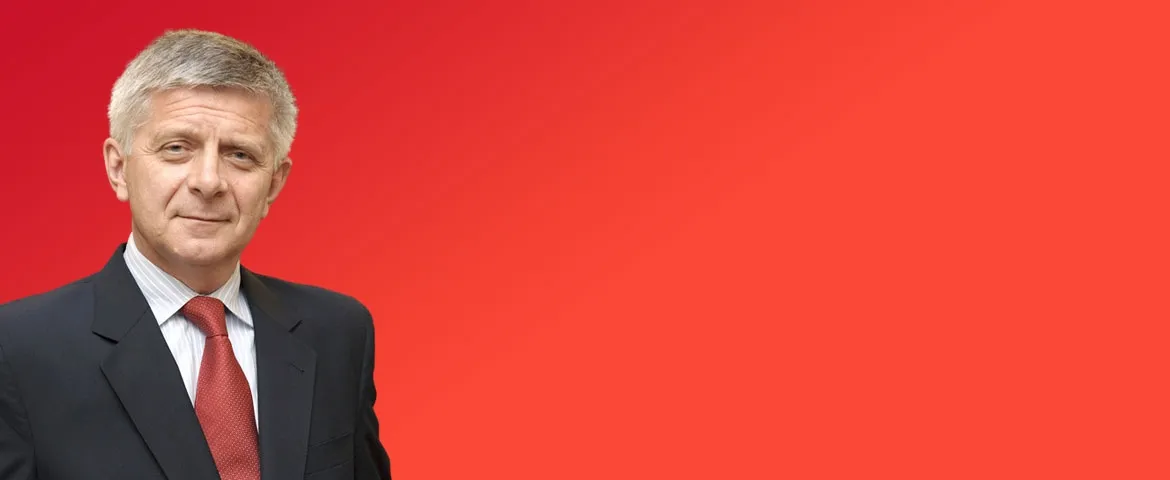 Mężczyzna w garniturze i czerwonym krawacie stoi na jednolitym czerwonym tle.