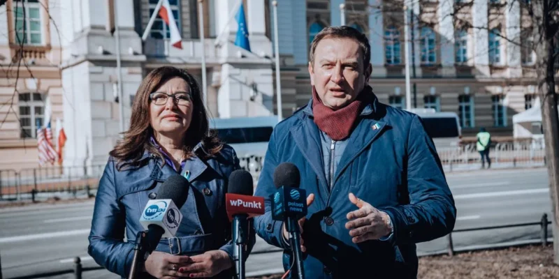 Na zdjęciu widać dwie osoby przemawiające do mediów przed budynkiem z flagami, przy stanowiskach reporterskich.