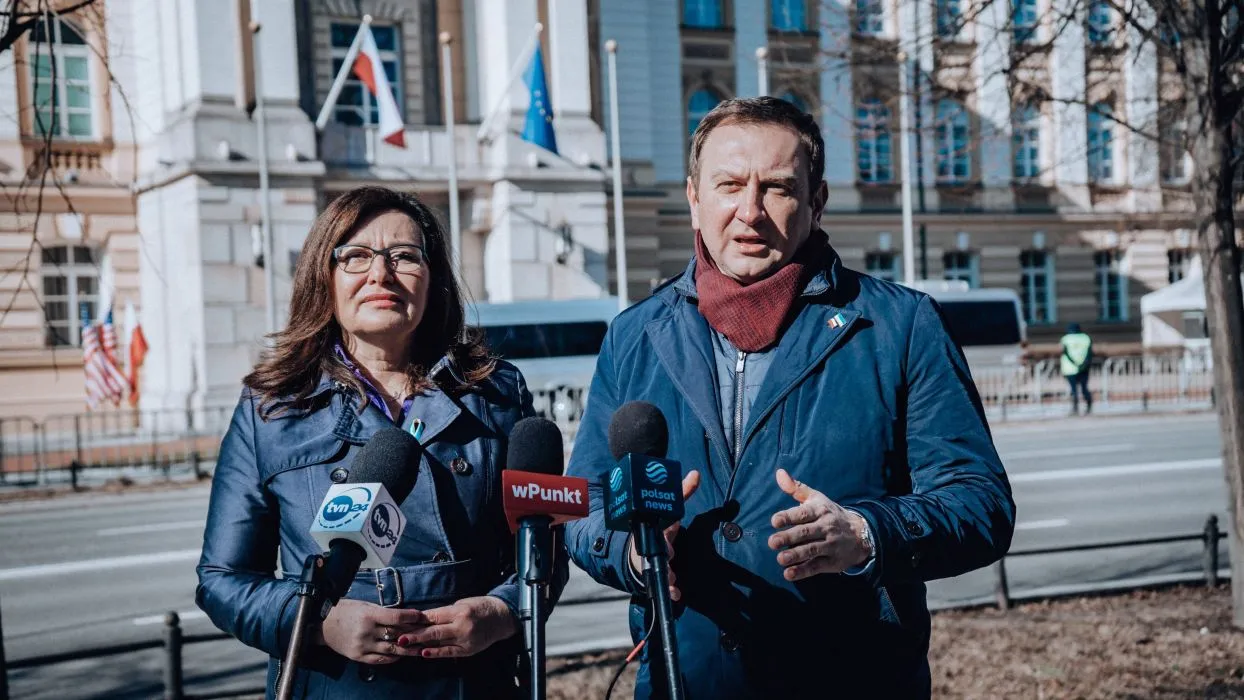 Na zdjęciu widać dwie osoby przemawiające do mediów przed budynkiem z flagami, przy stanowiskach reporterskich.