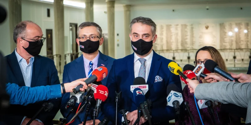 Na zdjęciu widać grupę mężczyzn i kobiet w garniturach, którzy udzielają wywiadu dziennikarzom biorącym udział w konferencji prasowej.