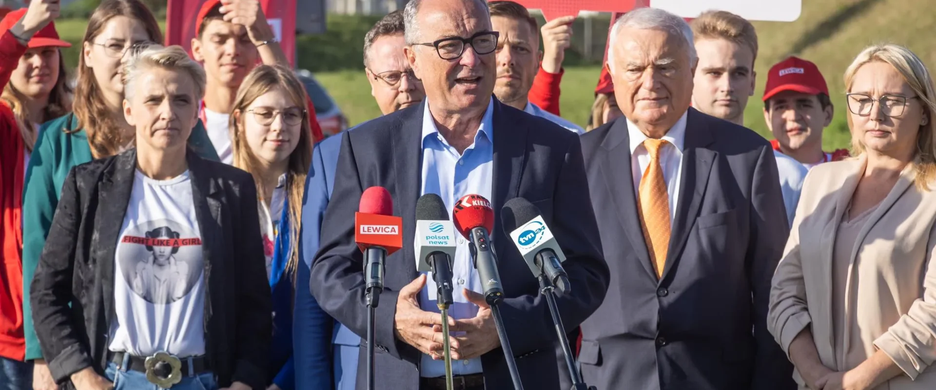 Mężczyzna w garniturze przemawia do zestawu mikrofonów podczas konferencji prasowej, otoczony przez zwolenników Lewicy i czerwone plakaty z napisem Lewica.