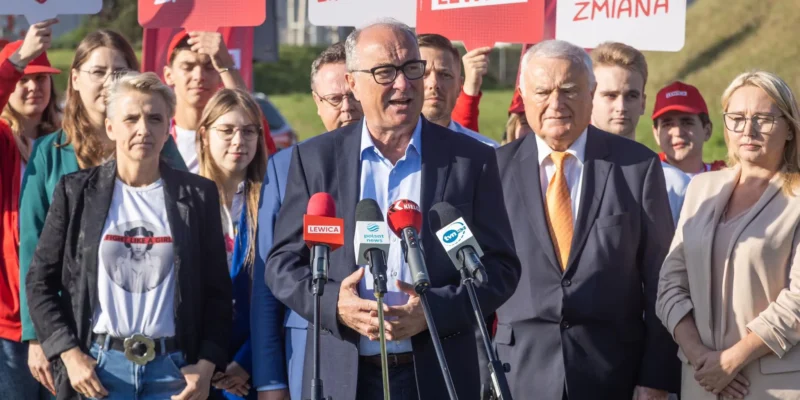Mężczyzna w garniturze przemawia do zestawu mikrofonów podczas konferencji prasowej, otoczony przez zwolenników Lewicy i czerwone plakaty z napisem Lewica.