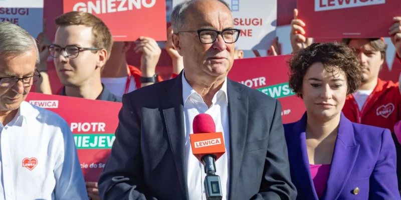 Mężczyzna w garniturze i okularach stoi przed mikrofonem z logiem Lewicy, otoczony przez zwolenników trzymających czerwone plakaty z hasłami Lewicy.