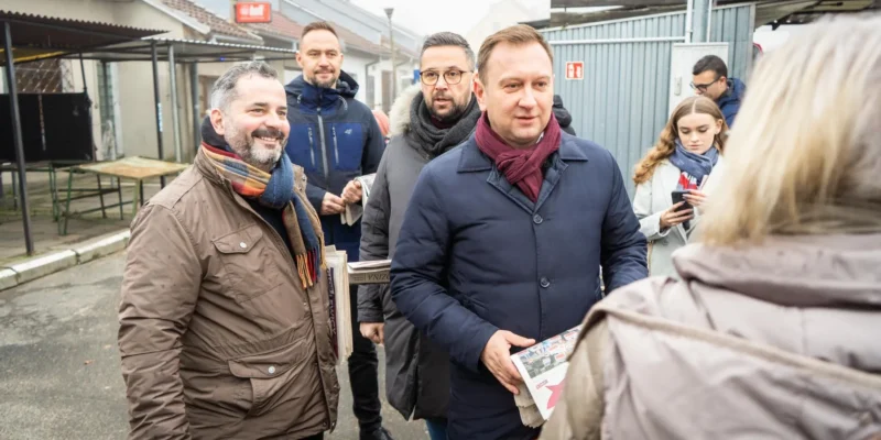 Grupa mężczyzn w ciepłych kurtkach i szalikach stoi na zewnątrz, w tle widać budynki oraz mgliste otoczenie, a jeden z mężczyzn trzyma prasę.